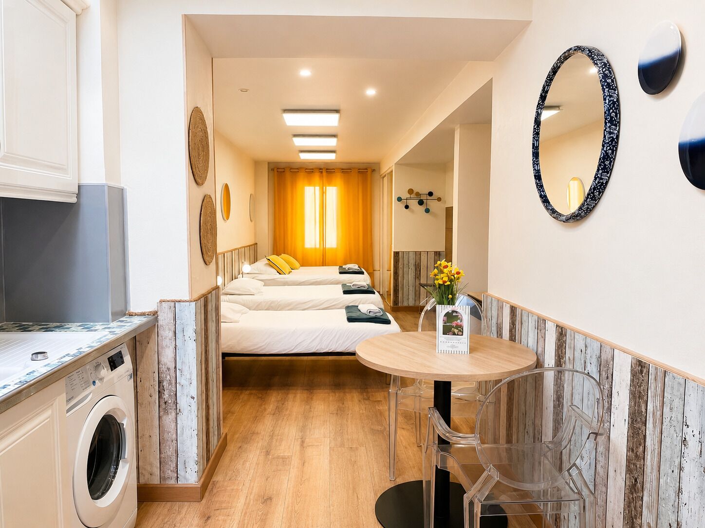 A hotel room with a washing machine, a bed with two pillows, a small dining table, and a mirror.