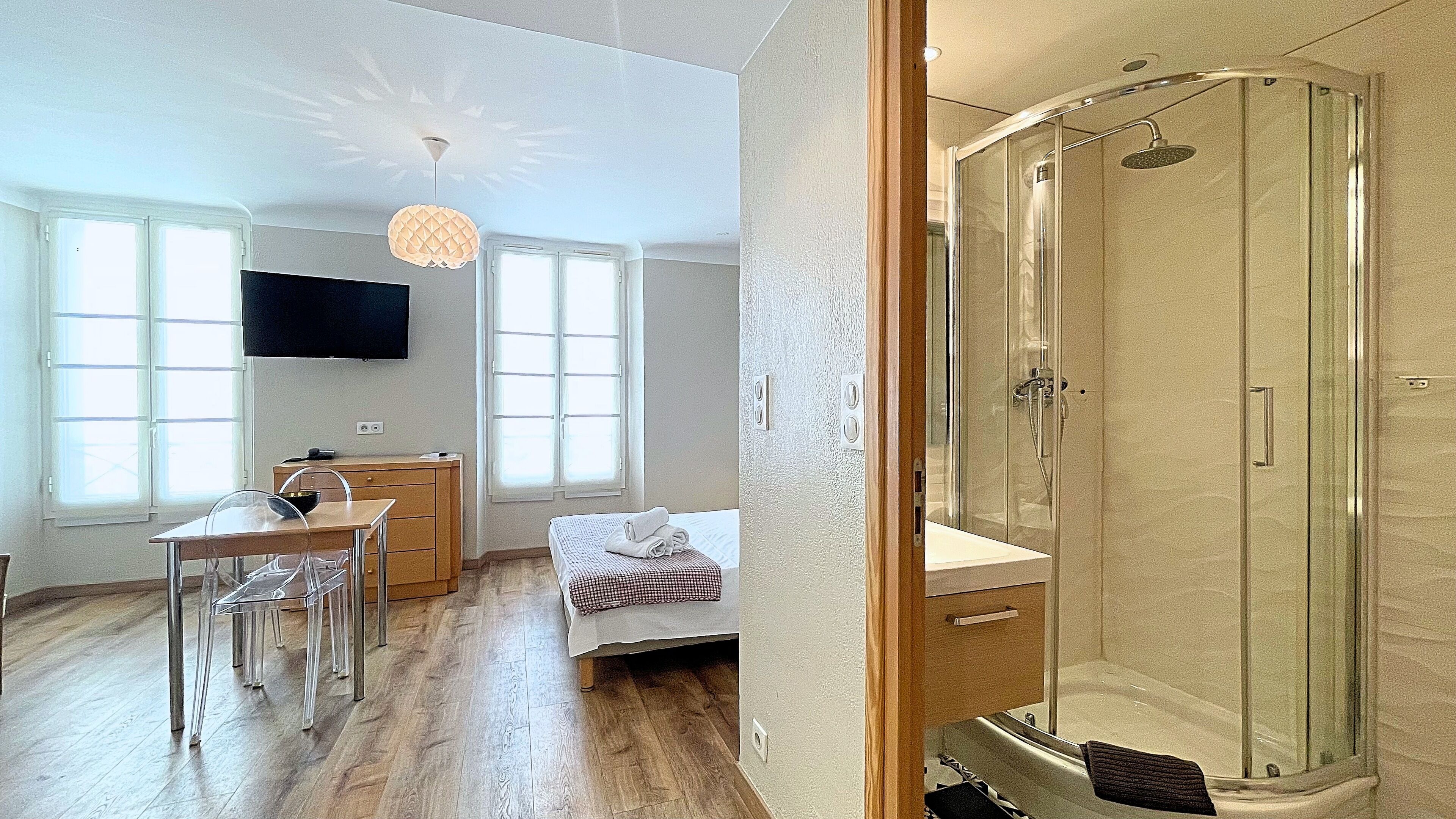 A modern hotel room with a wooden floor, a bed with a checkered blanket, a desk with a chair, a TV, and a glass-enclosed shower.