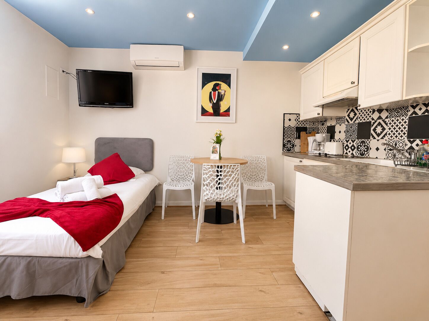 A hotel room with a bed, a TV, a small dining table with chairs, a kitchen area, and a ceiling light.