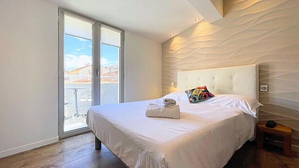 A modern bedroom with a large bed, a balcony with chairs, and a city view. - Hotel Le Florian (Cannes)