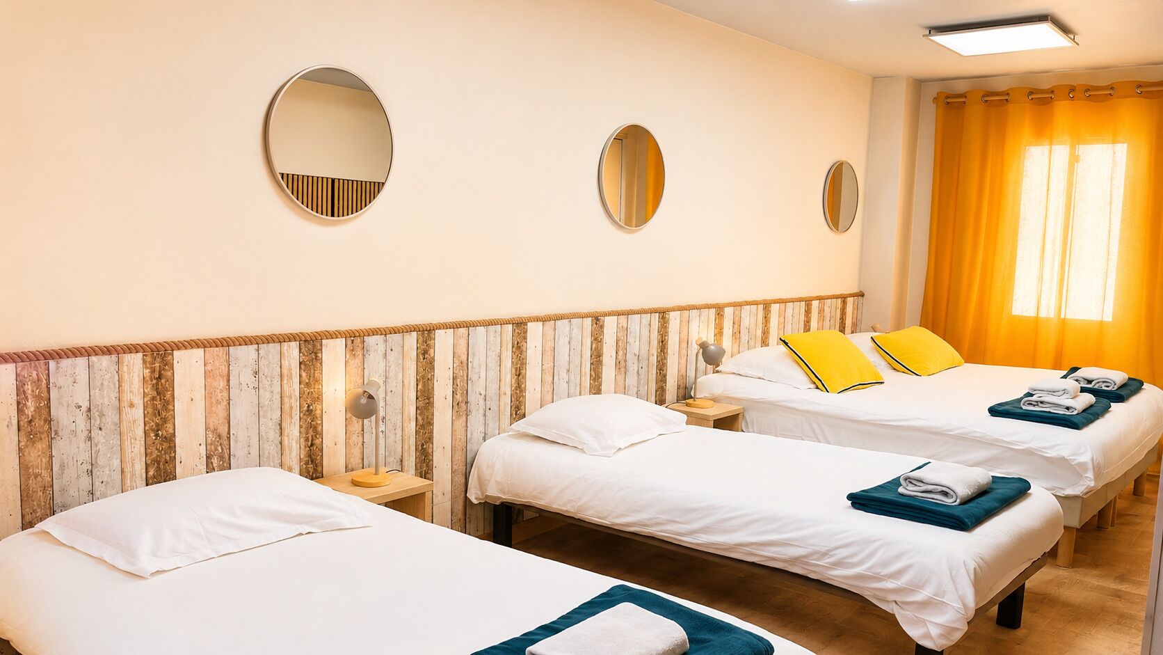 Ett hotellrum med tre enkelsängar, en sänggavel i trä, två sängbord med lampor och ett stort fönster med gardiner.