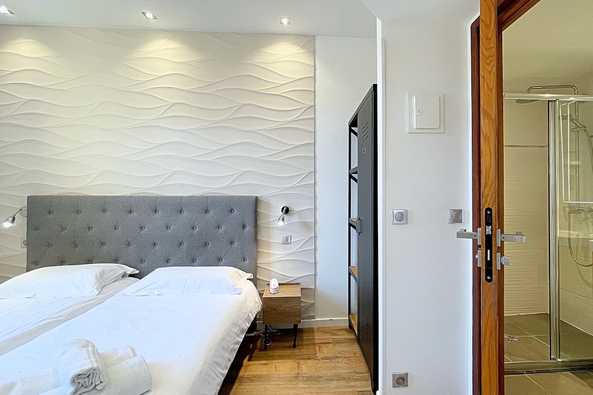A modern bedroom with a tufted headboard, a bed with white linens, a bedside table with a lamp, and a walk-in shower with a glass door.
