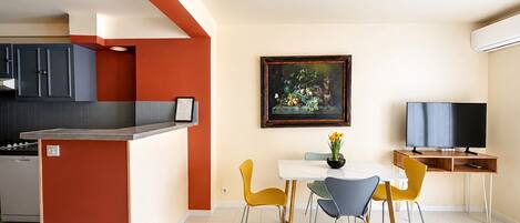 A modern kitchen with a red accent wall, a dining area with a white table and colorful chairs, and a painting on the wall.
