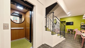 A modern interior with a wooden elevator, a staircase with black railings, a green wall, and a dining area with pink chairs. - Hotel Le Florian (Cannes)