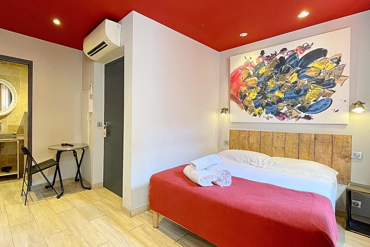 A modern hotel room with a large bed, a wooden headboard, a red accent wall, a large abstract painting, a round mirror, a small table, and a built-in air conditioning unit.