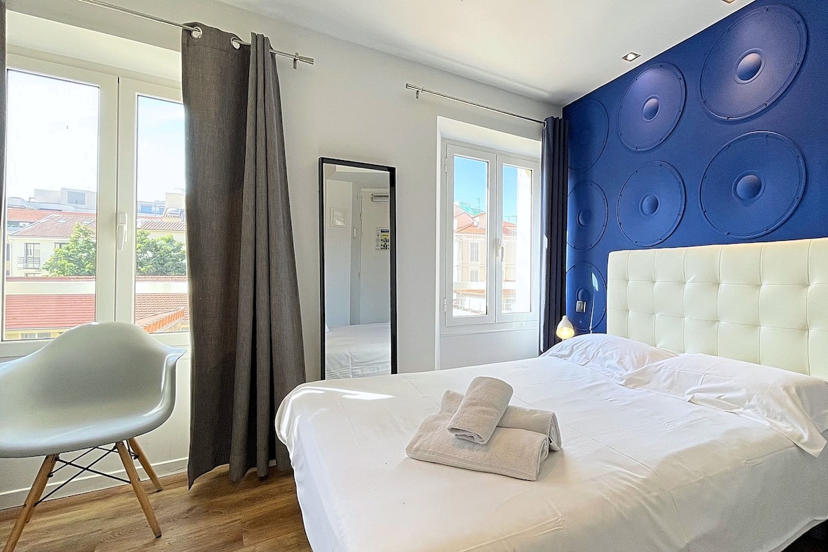 A hotel room with a bed, a chair, a bathroom door, and a view of buildings through the window.