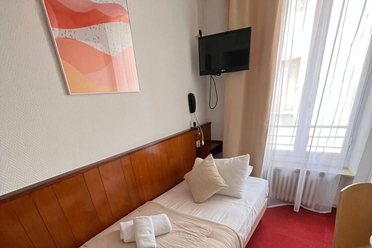 Habitación de hotel con cabecera de madera, cama con ropa blanca, alfombra roja, televisor montado en la pared y ventana con cortinas de gasa.