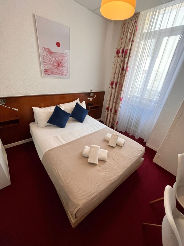 A hotel room with a bed, pillows, a headboard, a bedside table, a lamp, a window with curtains, and a wall art piece. - Azurene Royal Hotel (Cannes)
