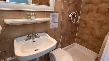 Un baño con lavamanos, inodoro y zona de ducha blancos. Hay un espejo sobre el lavamanos y una repisa con artículos de aseo.