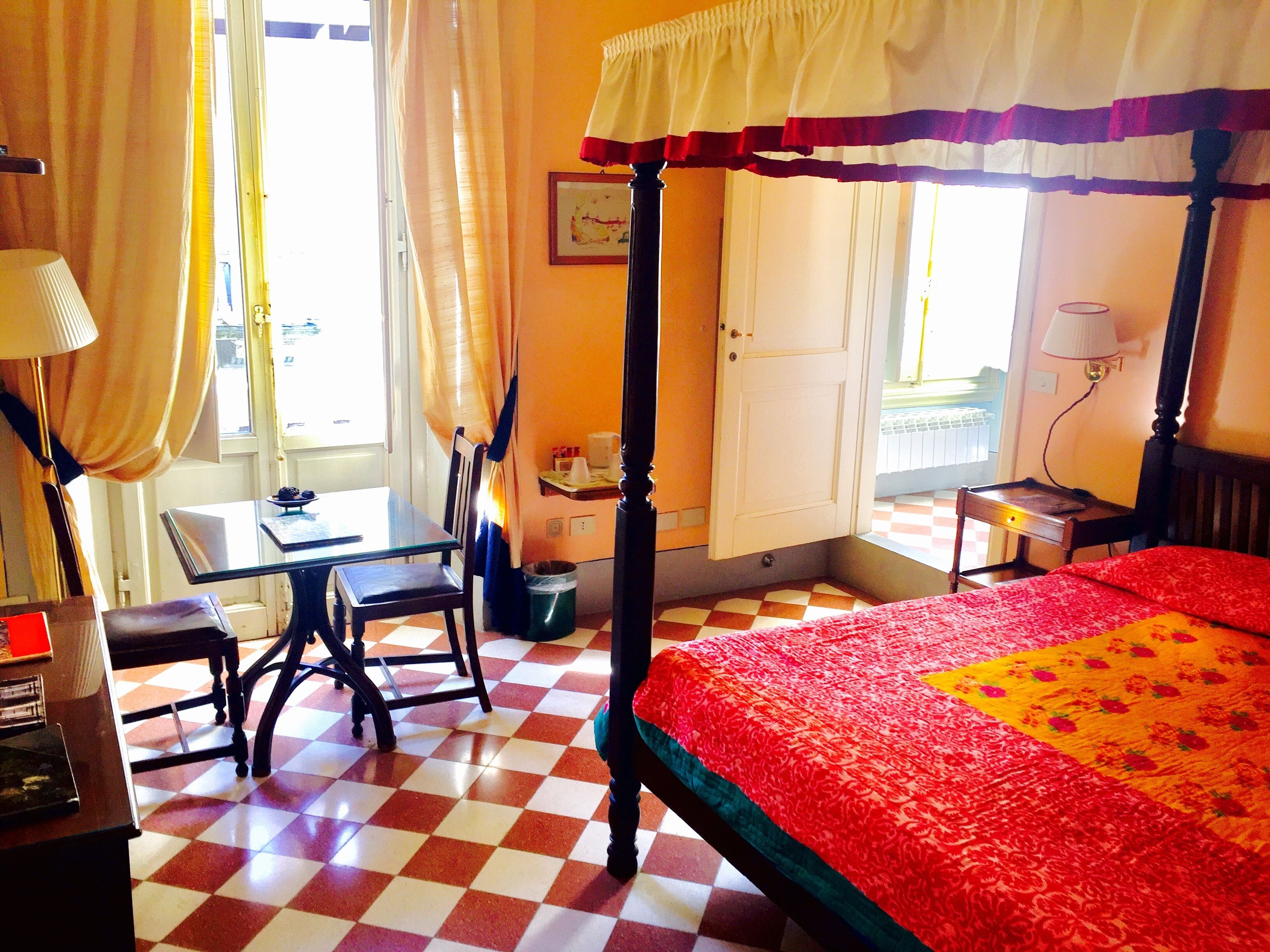 Ein Hotelzimmer mit Schachbrettmuster-Boden, einem Himmelbett, einem Schreibtisch mit Stuhl und einem Fenster mit Vorhängen.