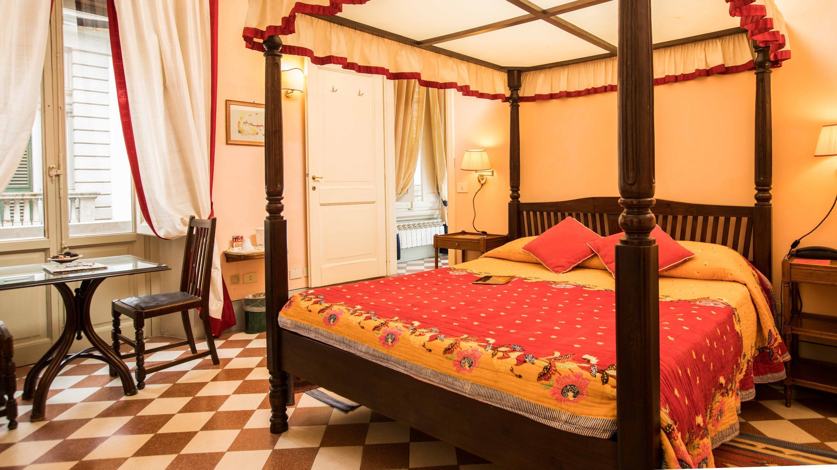 A four-poster bed with a red and yellow patterned bedspread, a wooden nightstand with a lamp, and a checkered floor.