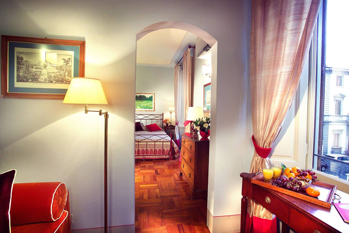 A hotel room with a wooden floor, a bed with a patterned bedspread, a dresser with fruit and a tray, and a view of the street through a window with curtains.