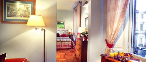 Habitación de hotel con piso de madera, cama con cubrecama estampado, cómoda con fruta y bandeja, y vista a la calle a través de una ventana con cortinas.
