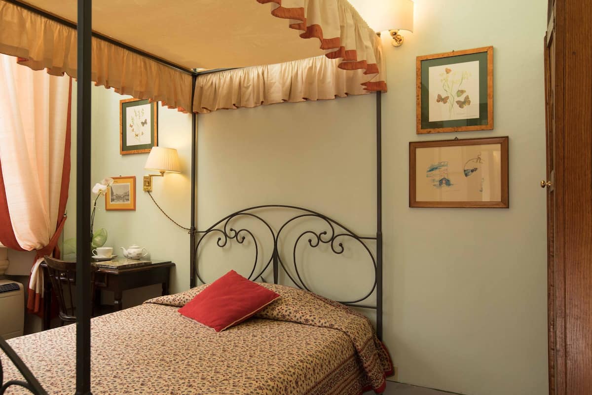 A four-poster bed with a floral bedspread and a red pillow, a bedside table with a lamp, and framed botanical prints on the wall.