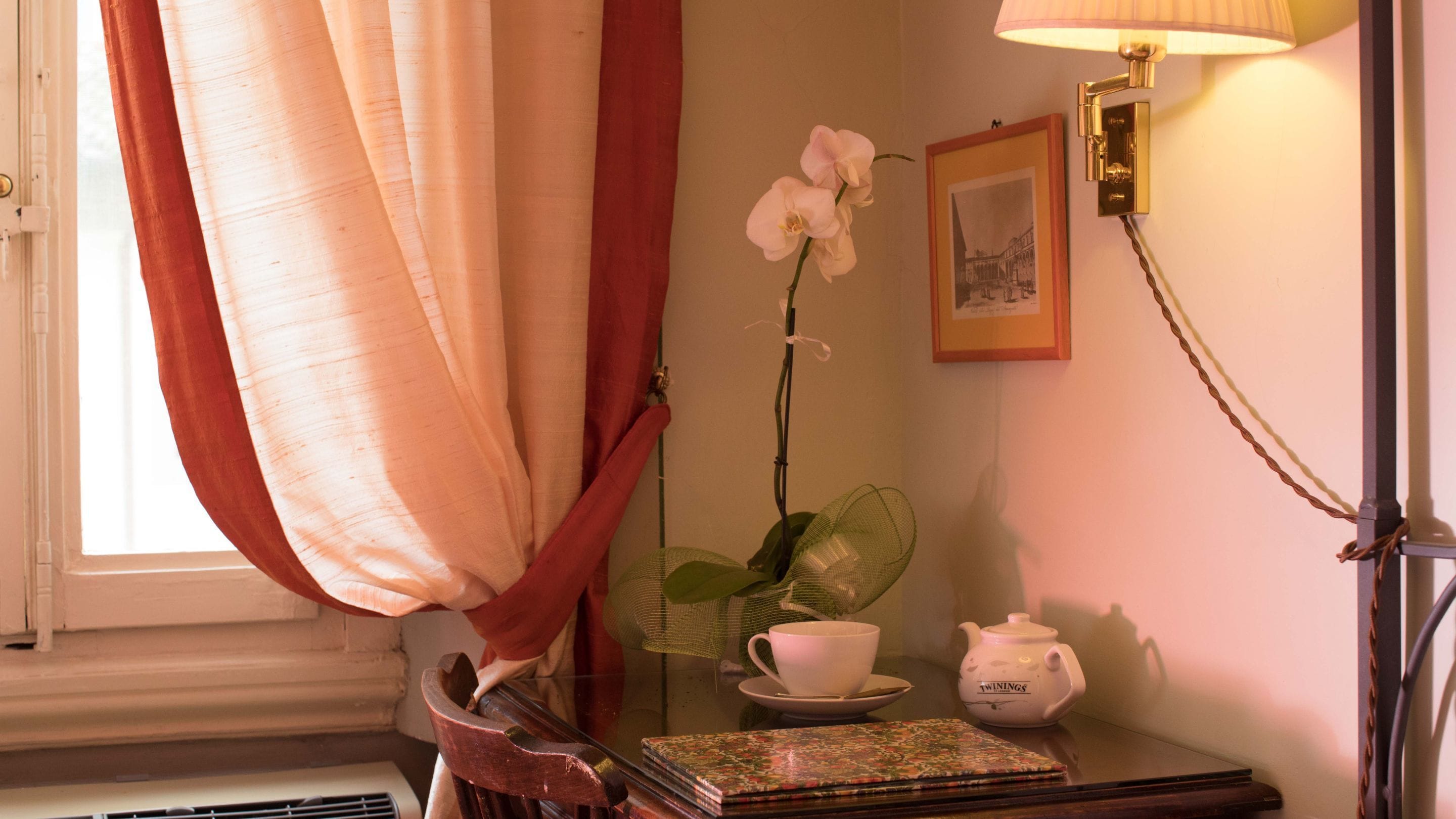 Une pièce avec une fenêtre, une table sur laquelle se trouvent une théière, une tasse, une soucoupe, un livre, une fleur et une lampe.