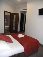 Ein Hotelzimmer mit einem Bett, einer rot-weißen Tagesdecke, einem weißen Kopfkissen und einem gefalteten Handtuch auf dem Bett. Eine Tür führt zu einem Badezimmer mit Toilette und Spiegel.