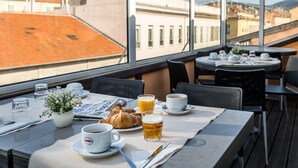 A dining table with breakfast items, a newspaper, and a view of buildings through large windows.