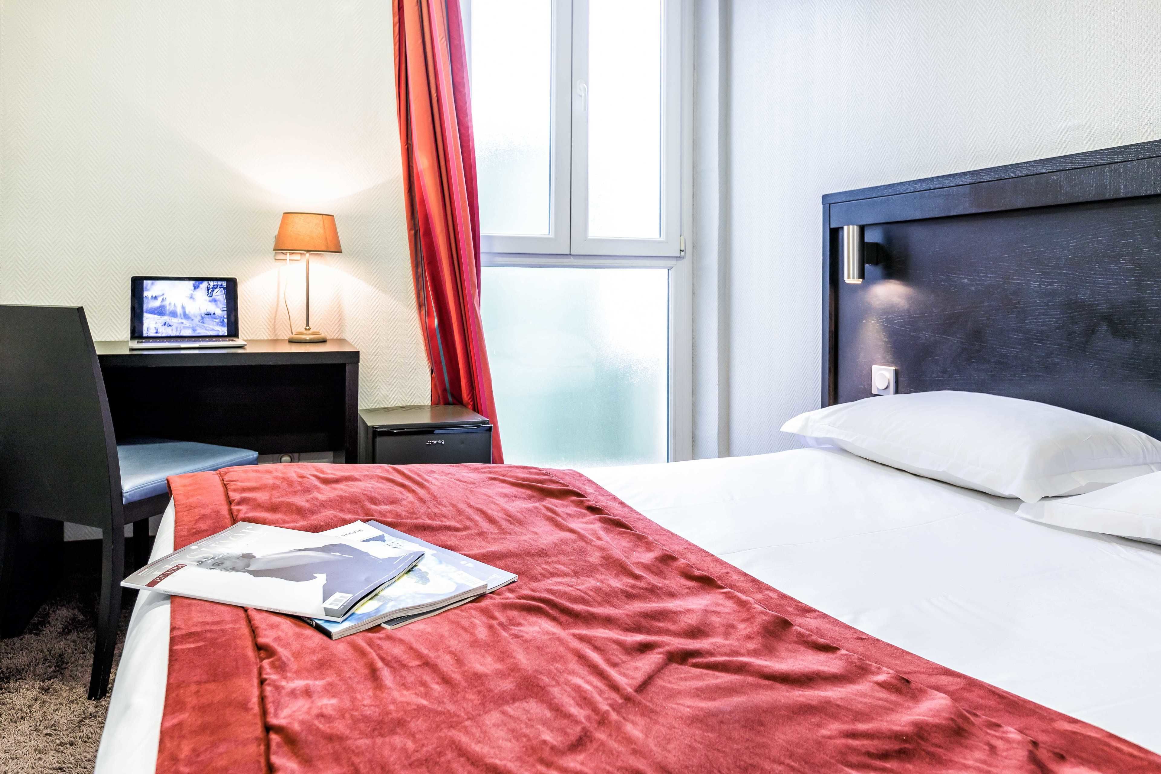 A hotel room with a bed, a desk with a laptop, a lamp, and a window with curtains.
