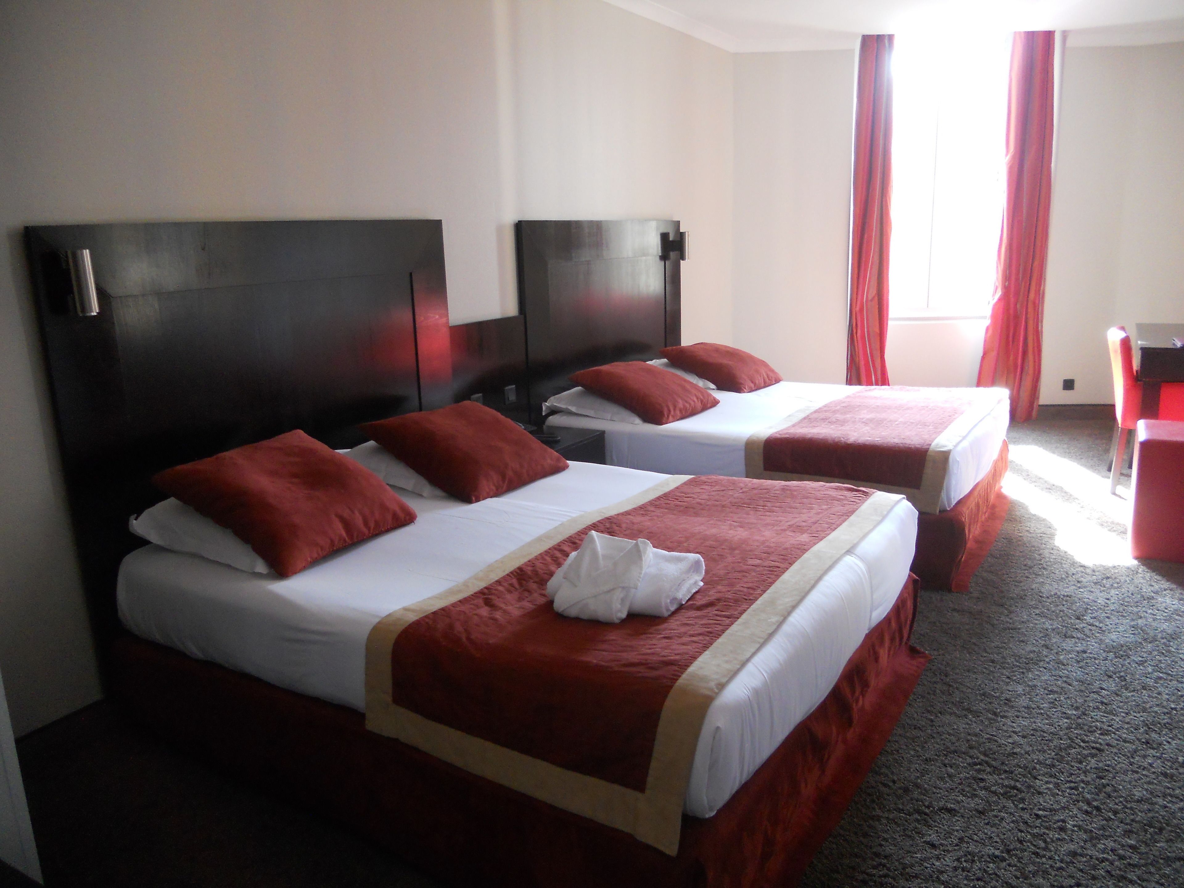 Una camera d'albergo con due letti, ognuno con lenzuola rosse e biancheria bianca, una testiera in legno e una sedia rossa.