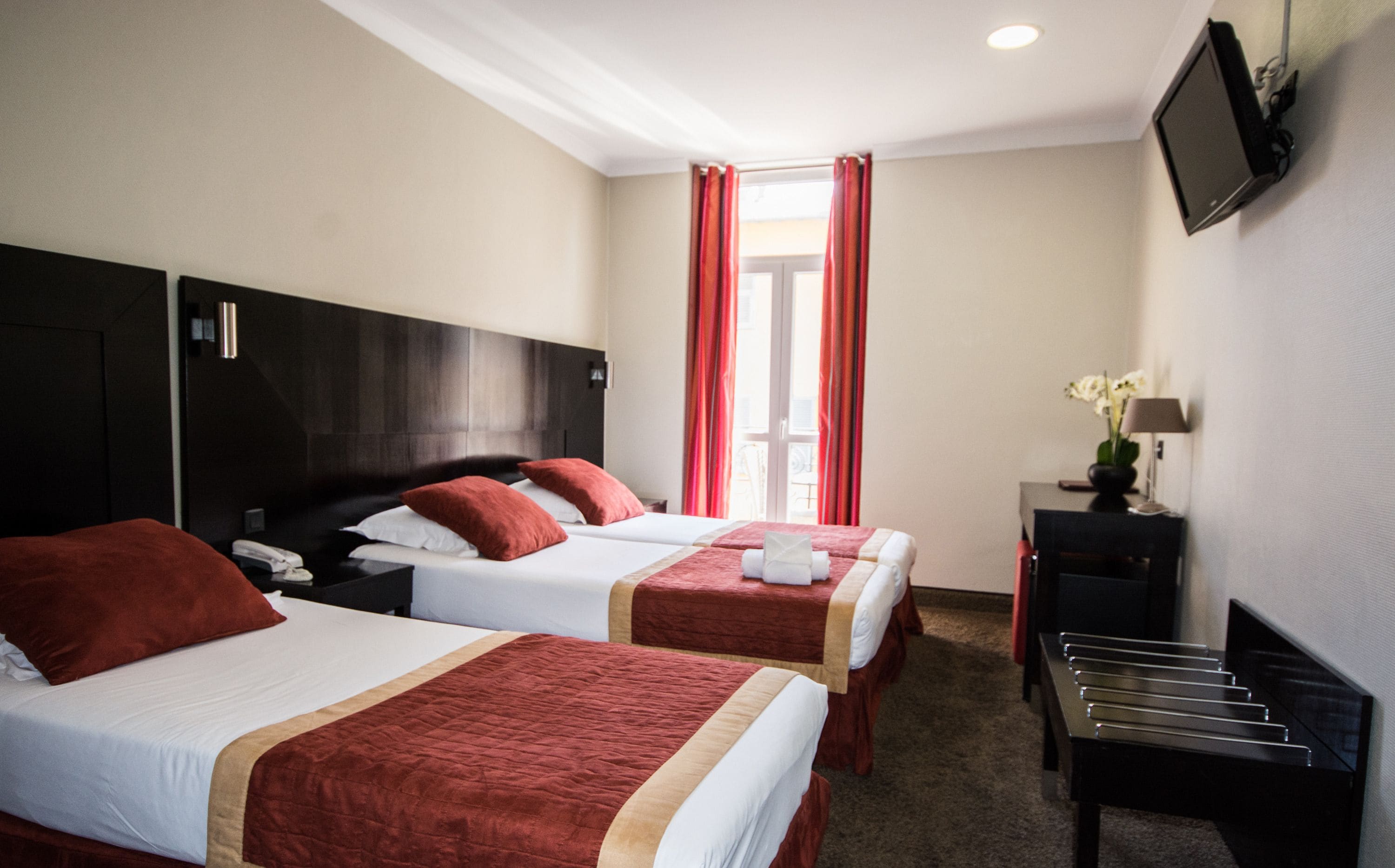 A hotel room with two beds, a desk, a TV, and a window with red curtains.