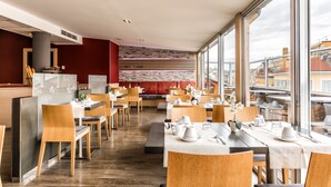 A modern restaurant with wooden furniture, white tablecloths, and a view of the city through large windows.