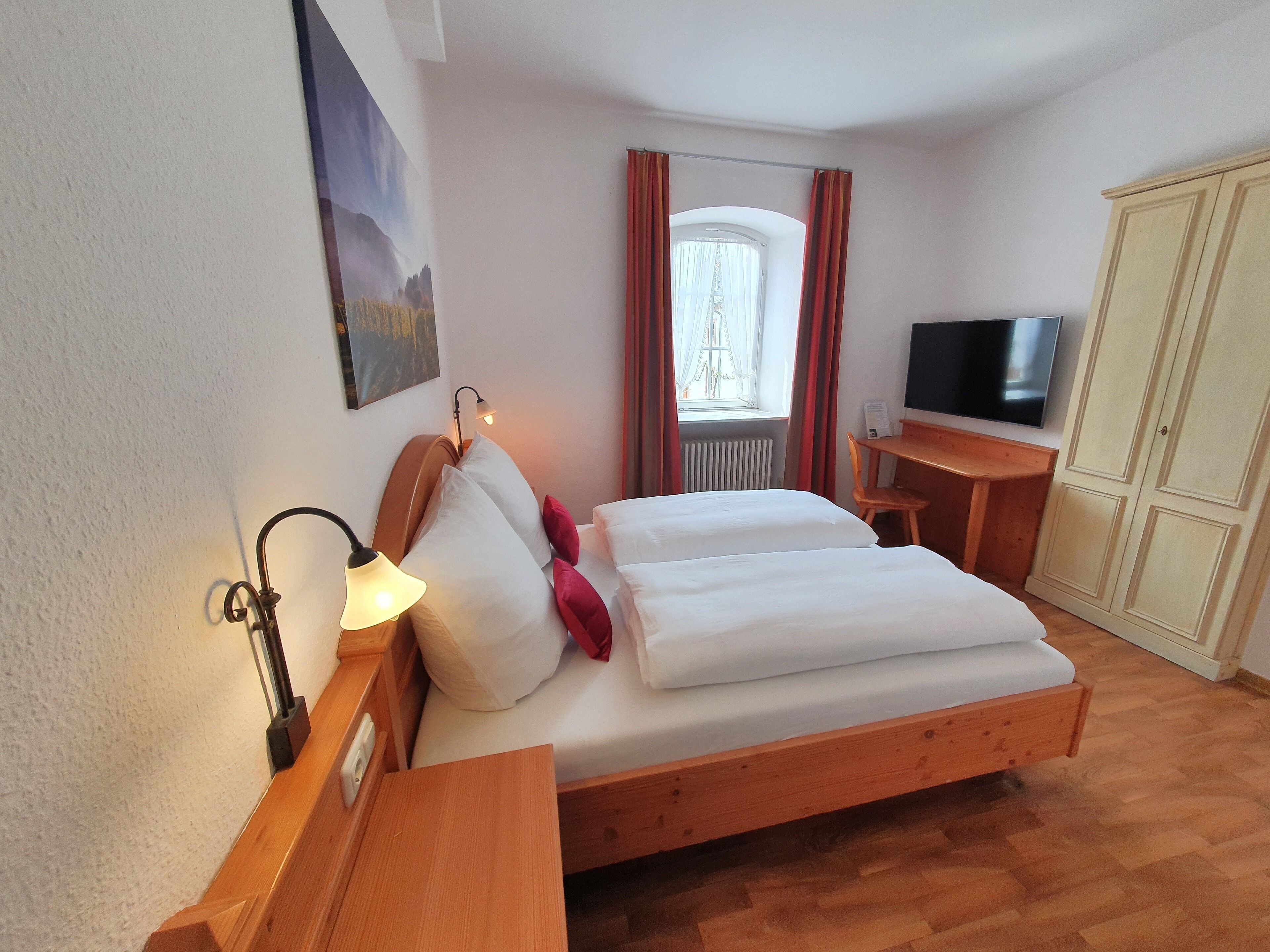 A bedroom with a bed, bedside lamp, a desk with a chair, a television, and a wardrobe.