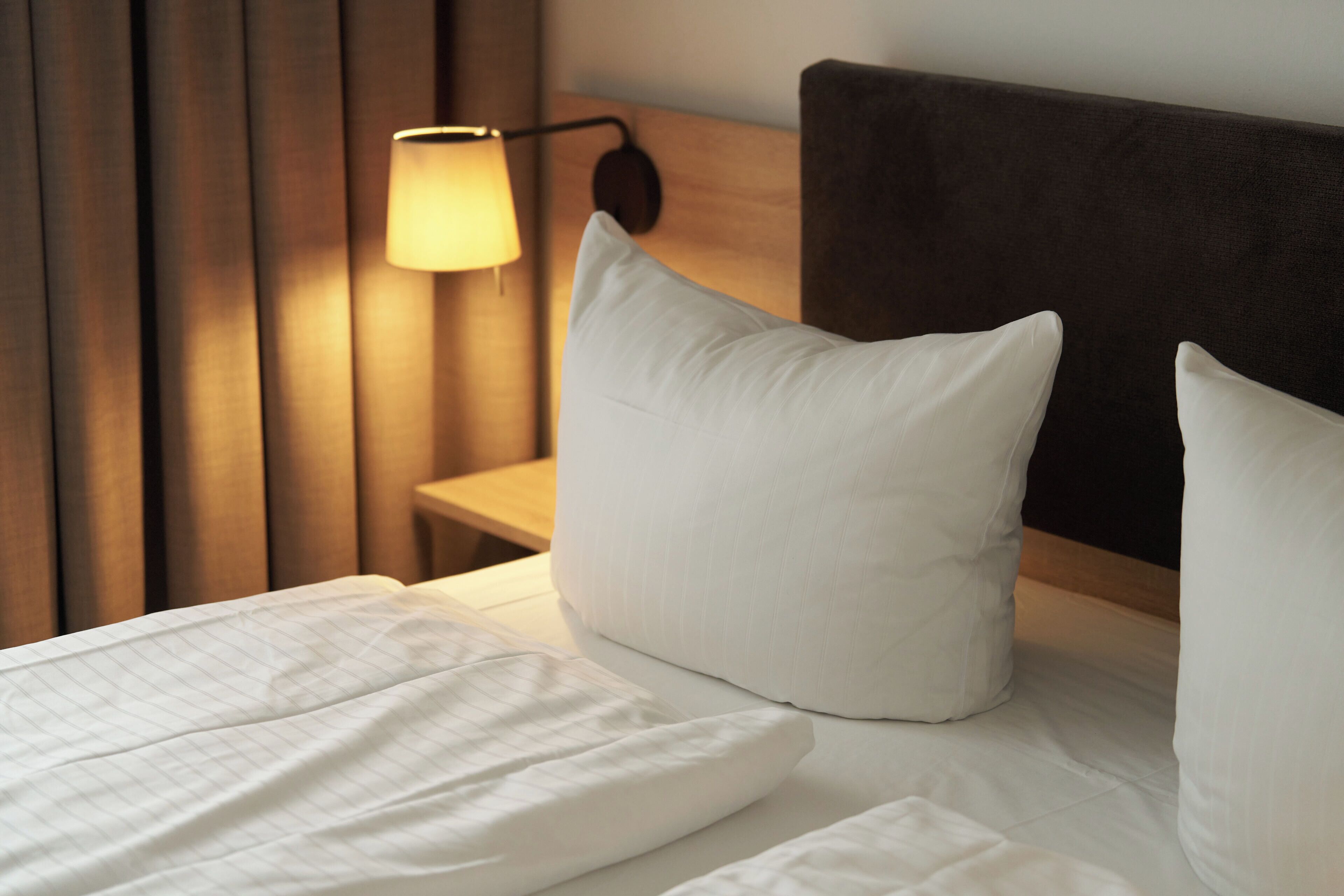 A neatly made bed with white linens, a wooden headboard, and a wall-mounted lamp.