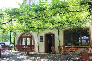 An outdoor cafe with tables and chairs under a tree, a sign reading "Frische Hack-Steaks," and a building entrance.