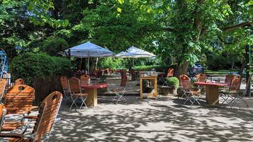 Een terras met tafels en stoelen, parasols en bomen die schaduw bieden.
