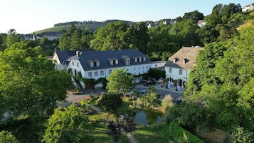 A large, white building with multiple windows and a sloped roof, surrounded by lush greenery and a small pond.