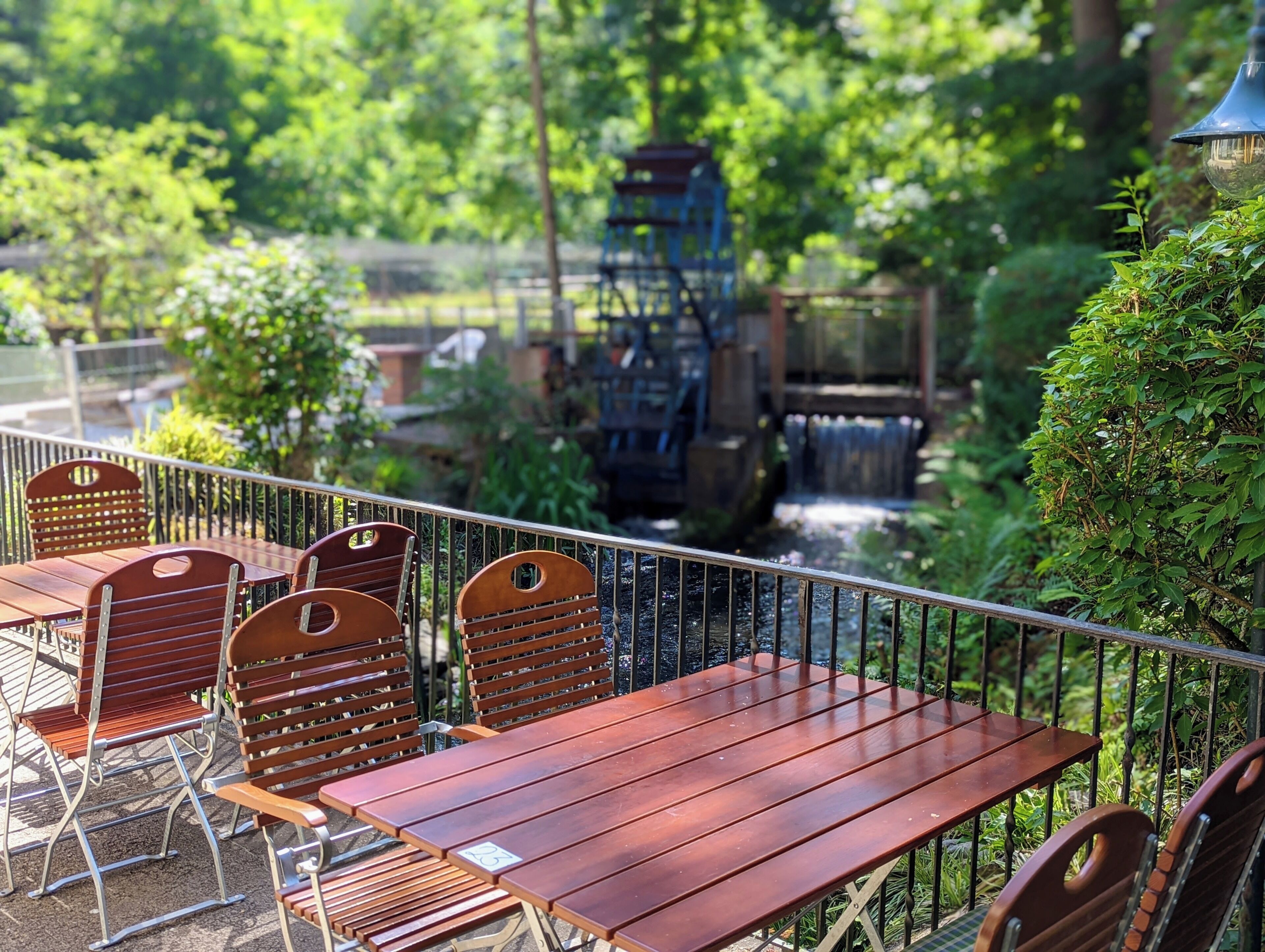 An outdoor seating area with wooden tables and chairs, surrounded by greenery and a water feature.