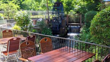 An outdoor seating area with wooden tables and chairs, surrounded by greenery and a water feature.