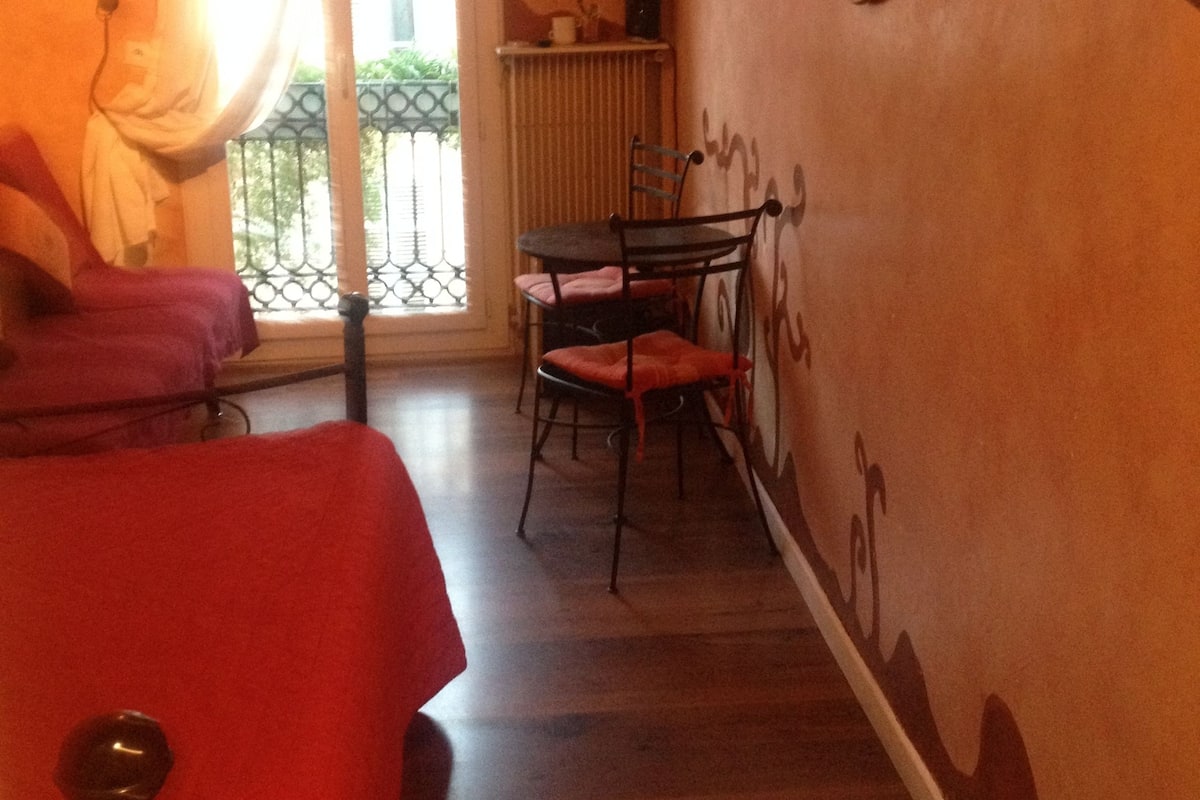 A hotel room with a bed, a chair, a table, a TV mounted on the wall, and a window with curtains.