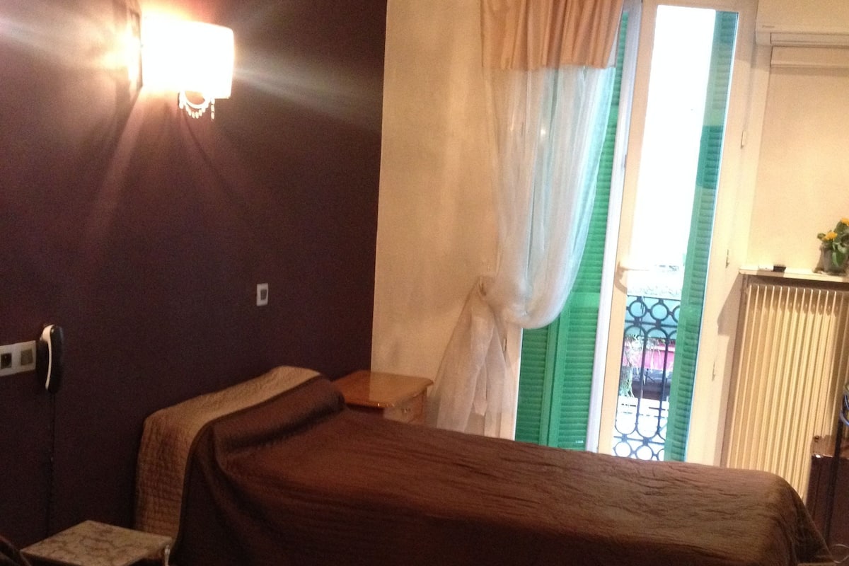 A hotel room with two beds, a small table, a chair, a window with curtains, and a radiator.