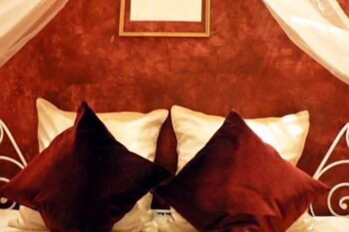 A bed with white metal frame and dark red pillows, a framed picture on the wall, and sheer curtains.