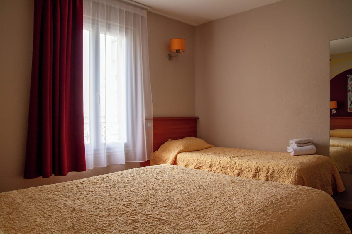 Quarto de hotel com duas camas, janela com cortinas leves, luminária na parede e uma toalha dobrada na cama.