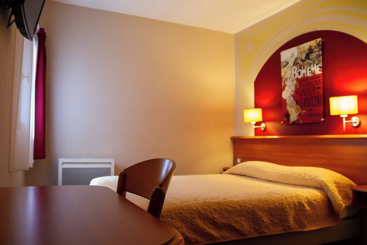 Quarto de hotel com uma cama, uma mesinha de cabeceira de madeira com abajures, uma cadeira e um quadro na parede.