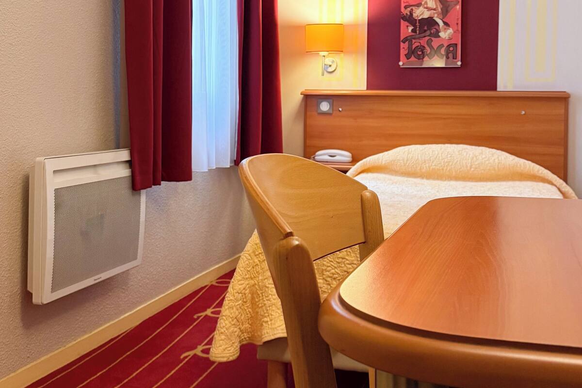 Quarto de hotel com cama, uma cadeira de madeira, uma mesa, uma luminária, um cartaz na parede e uma janela com cortinas.