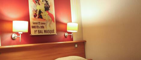 Une chambre d’hôtel avec un lit, deux lampes de chevet, une table de chevet, une affiche au mur et un mur rouge.