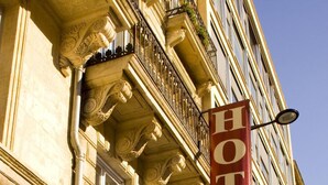 La façade d’un hôtel est ornée d’une grande banderole rouge sur laquelle on peut lire « HÔTEL », et d’une enseigne indiquant « CHEZ GREG LE GRAND THÉÂTRE » en dessous.