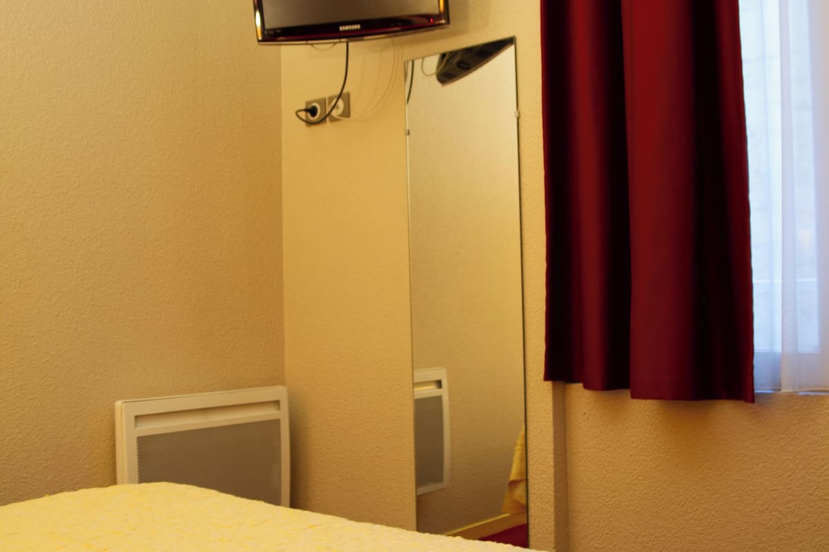 Quarto de hotel com cama, televisão na parede, espelho e janela com cortinas.