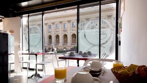 Une table mise pour le petit-déjeuner, avec des chaises, et une vue sur un bâtiment historique par de grandes fenêtres.