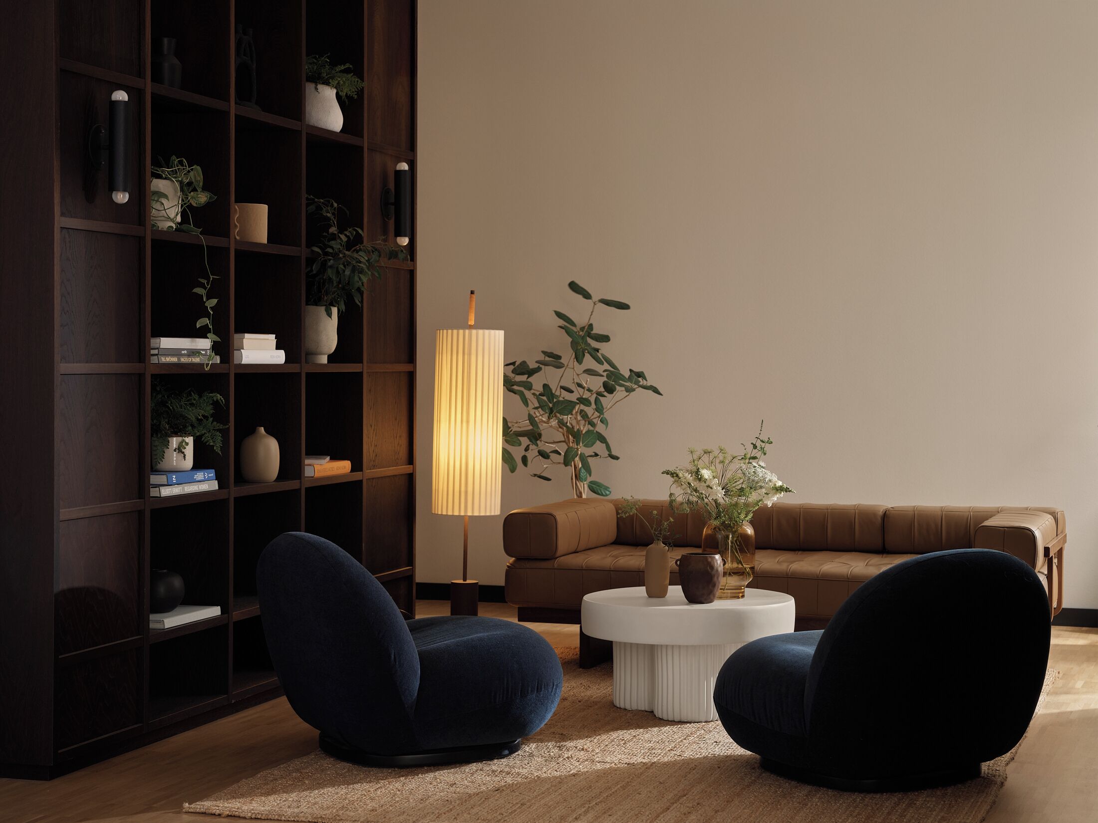 Un salon moderne avec une bibliothèque en bois foncé, deux fauteuils bleu foncé, une table basse ronde blanche et un grand lampadaire.
