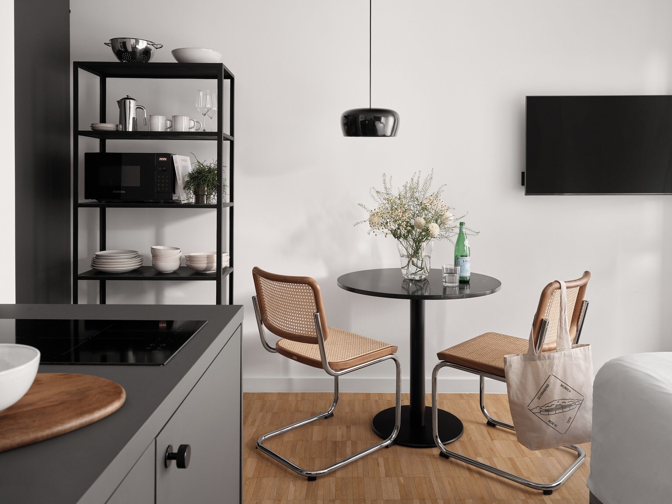 A modern kitchen with a dining area, featuring a black-framed table, two wicker chairs, a potted plant, and a bottle of water.