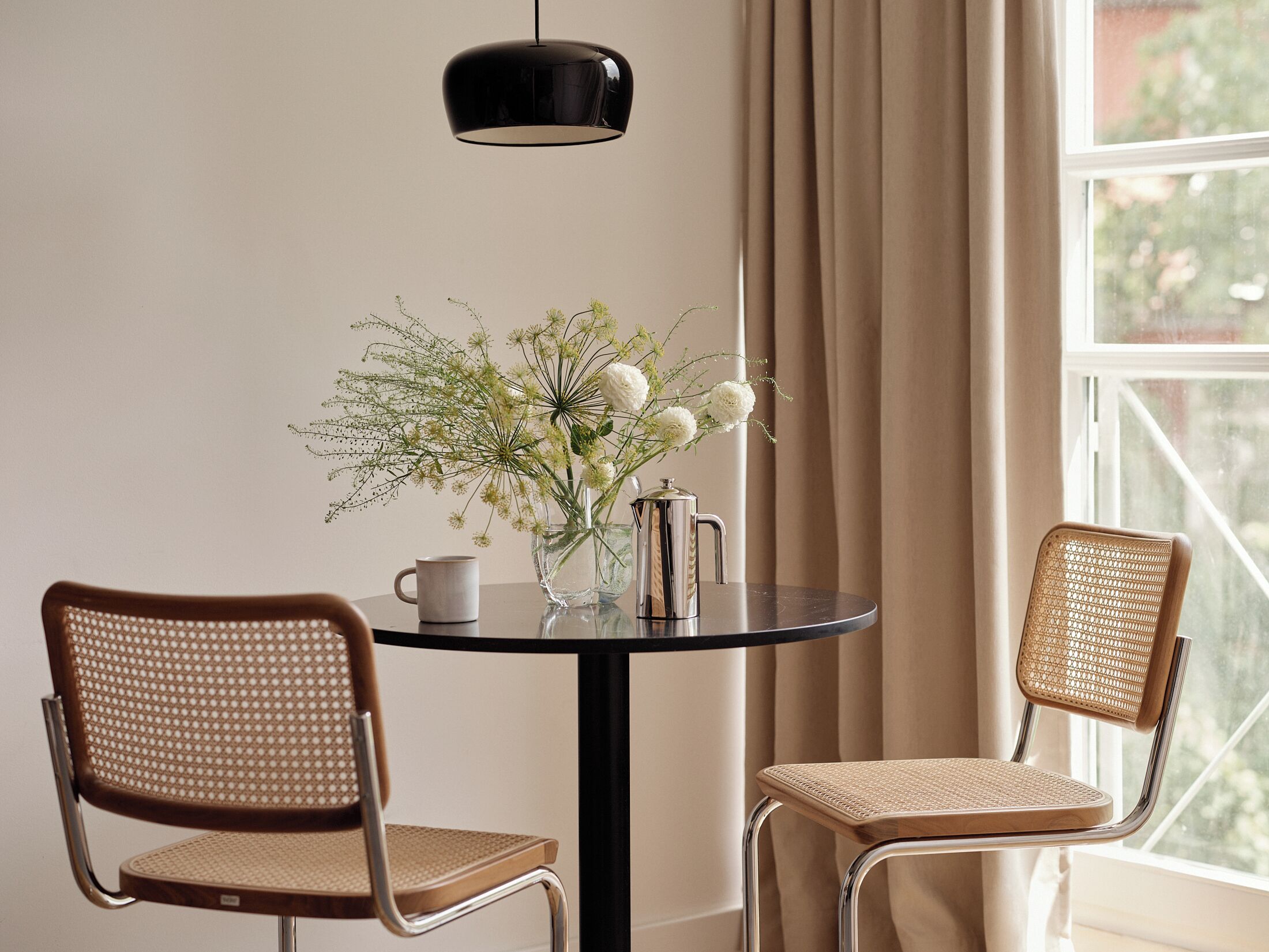 A round table with two wicker chairs, a vase with flowers, a coffee pot, and a black pendant light.