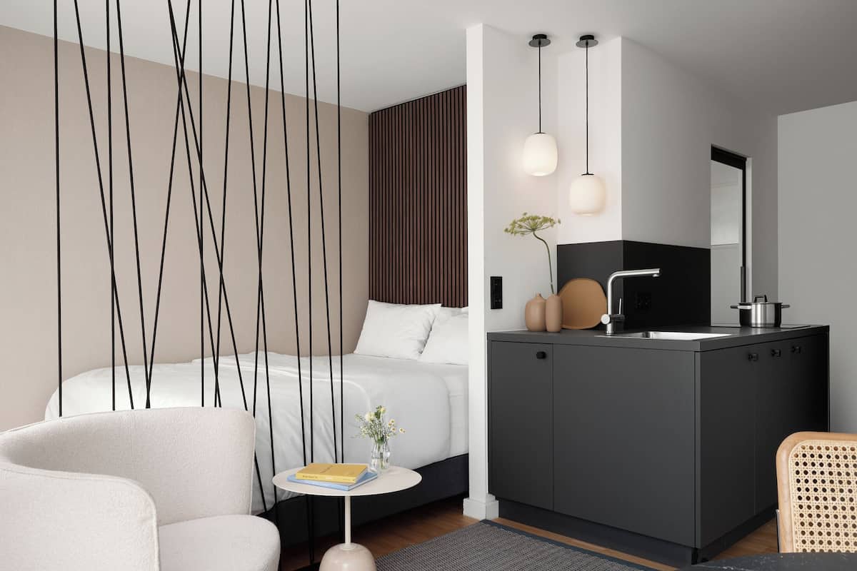 Une chambre d’hôtel moderne comprenant un lit, une petite table ronde avec un livre, un canapé, un coin cuisine avec un évier et des placards, et un grand miroir.