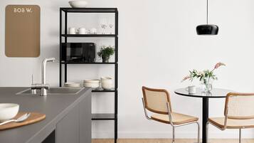 A modern kitchen with a built-in microwave, a black shelving unit with dishes and a plant, and a dining area with two wicker chairs and a table.