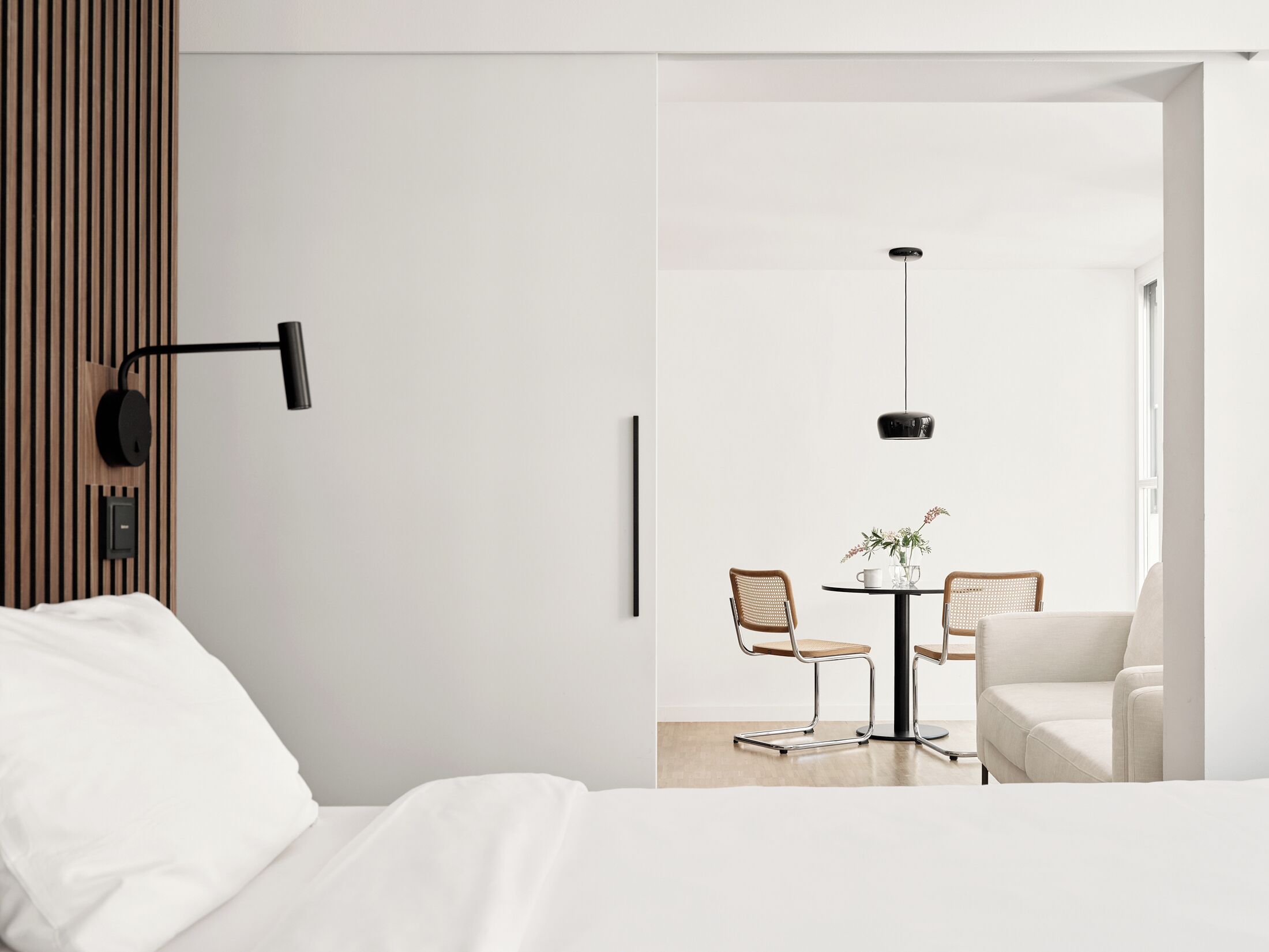 A modern hotel room with a bed, a wall-mounted lamp, a dining area with two chairs and a table, and a hanging pendant light.