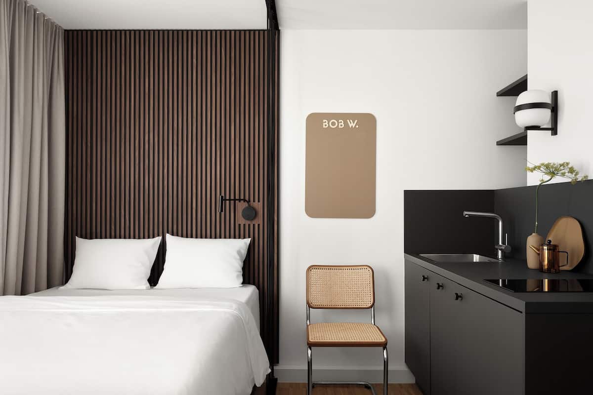 Une chambre d’hôtel moderne équipée d’un lit, d’une chaise, d’un bureau avec un lavabo et d’un luminaire fixé au mur.
