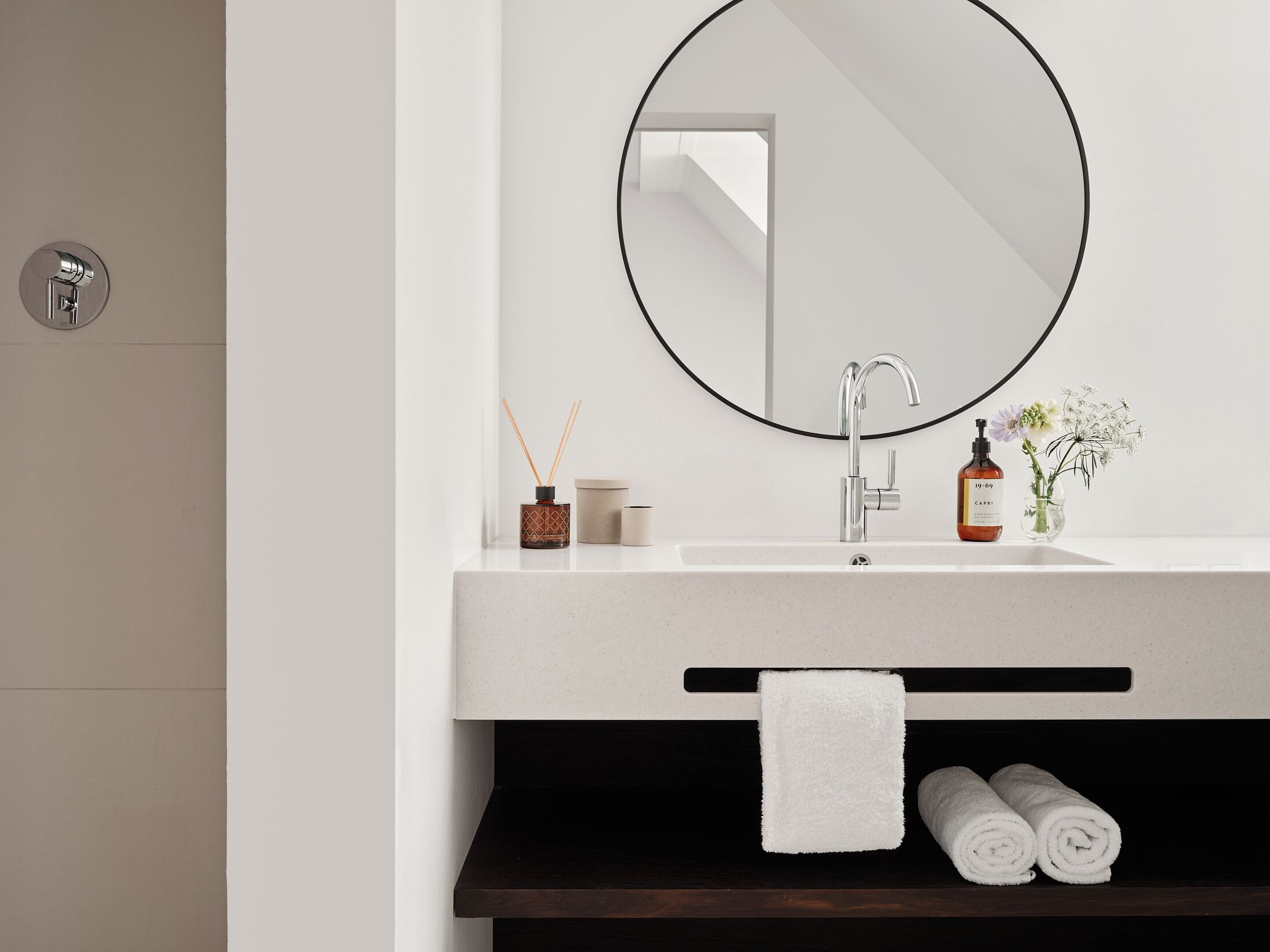 Ein modernes Badezimmer mit einem weißen Waschbecken, einem runden Spiegel und einem dunklen Holzregal darunter.